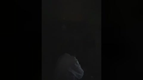 A video of a dimly lit room with a person in the background, partially obscured by shadow. The person is wearing a dark jacket and appears to be standing near a window. The image is dark and grainy.