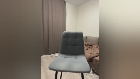 Video of a minimalist bedroom with a grey upholstered chair, beige sheets, brown curtains, and a white wall.
