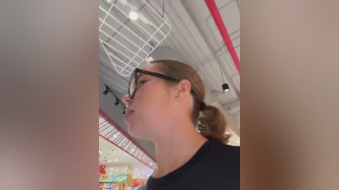 Media: A video of a young woman with light skin and brown hair in a ponytail, wearing glasses, black shirt, and standing in a colorful, modern, indoor setting with a high ceiling and red accents.