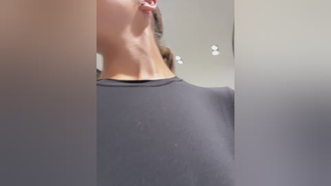 Media: Video of a person's neck and shoulder, covered in a gray, long-sleeved garment, likely a medical gown, in a dimly lit, sterile room with overhead lights and blurred background.