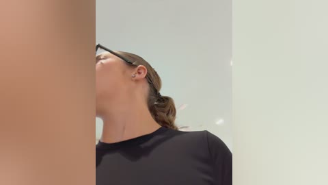 Media: Video of a woman with light skin, brown hair tied back, wearing black glasses and a black shirt, looking upward with an expression of surprise. The background is a plain, light-colored wall.