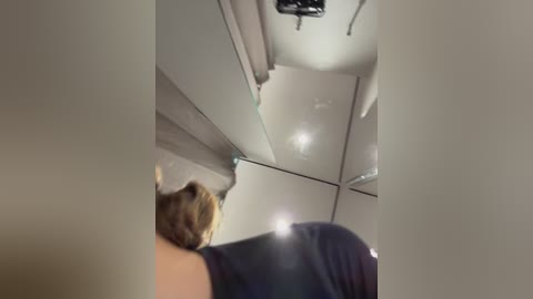 Media: A blurry video of an airplane's interior shows a person with brown hair, wearing a black shirt, looking down. The ceiling features bright overhead lights and a vent.