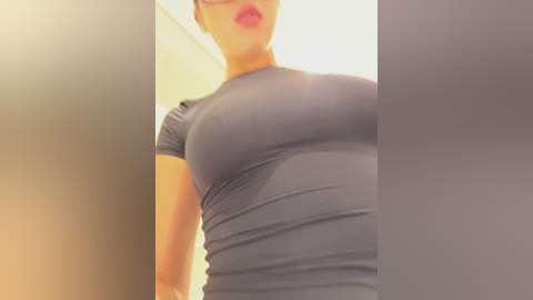 Media: Video of a light-skinned woman with medium-sized breasts wearing a tight, grey athletic top, her lips painted pink, against a blurred indoor background.