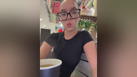 Media: A video of a woman with glasses and a black shirt sitting in a cafeteria, holding a white coffee cup, with blurred faces in the foreground.