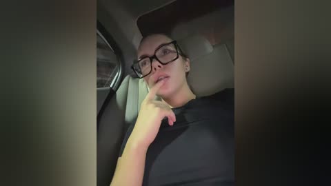Media: A video of a young woman with light skin, dark hair tied back, and glasses, resting her chin on her hand, sitting in a car.