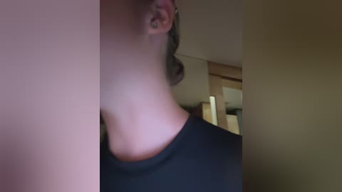Media: Video of a woman with light skin and shoulder-length dark hair, wearing a black t-shirt, seen from the side. Background shows a blurred indoor setting with neutral-colored walls and furniture.