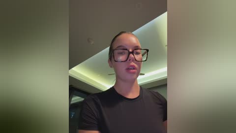 Media: Video of a young woman with fair skin, dark hair pulled back, wearing black-rimmed glasses and a black t-shirt, standing in a modern, dimly lit room with green ceiling lights.