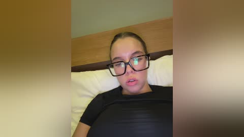 Media: Video of a young Caucasian woman with light skin, brown hair in a bun, wearing black-framed glasses and a black T-shirt, lying on a bed with white pillows, wooden headboard, and a blurred background.