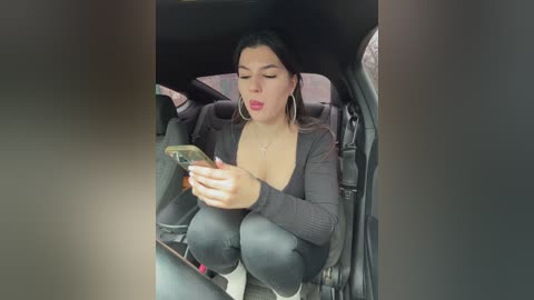 Media: Video of a young Latina woman with medium skin tone and dark hair, wearing a black cardigan, black leggings, and hoop earrings, sitting in a car, intensely texting.