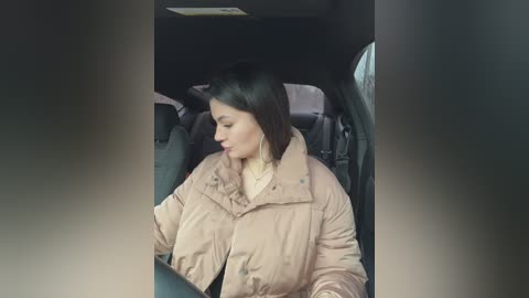 Media: Video of a woman with straight, dark hair and light skin, wearing a beige puffer jacket, seated in a car with black interior. She is gazing out the window, appearing contemplative.