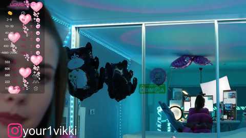 Media: Video of a room with a woman in a pink top, heart emojis, and stuffed animals on a white wall, with a reflection of a person in a purple shirt in the background.