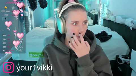 Media: Video of a young woman with fair skin, light brown hair, wearing a gray hoodie and large headphones, playfully covering her mouth, in a cozy, dimly-lit bedroom with a bed, stuffed animals, and a green \"YourVikki\" watermark.