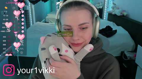 Media: A video of a young woman with light skin and long brown hair, wearing headphones, smiling while hugging a stuffed bear. Background shows a cluttered bedroom with fairy lights and a bed. Overlay includes social media icons.