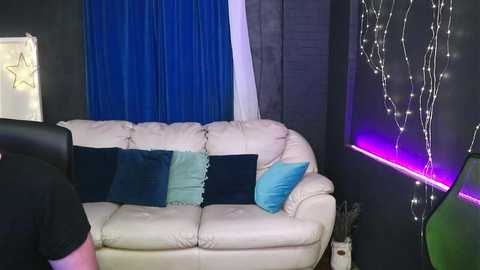 Video of a modern, dimly-lit living room with a white leather couch, blue and teal pillows, dark walls, and string lights.