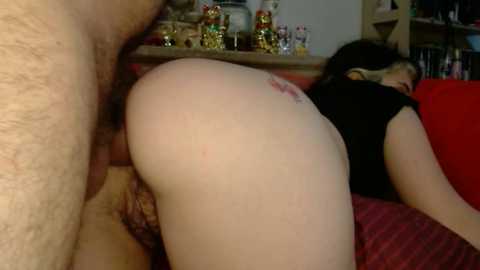A video captures a close-up of a man's erect penis penetrating a woman's anus while she lies face down on a red couch, wearing a black shirt.