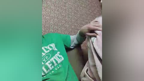Media: A video of a person in a green shirt with white text reading \"YOUTH\" and \"LEAGUE\" partially visible. They are holding a pink garment against a patterned, pink wall. The image has a green gradient overlay.