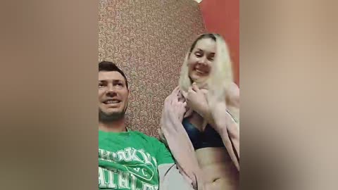 Media: A video features a smiling couple in a dimly lit room. The man, in a green shirt, and woman, in a black bra and pink robe, stand against a patterned wall.