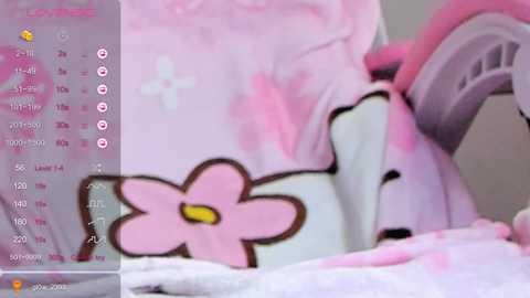 A close-up video of a pink and white plush animal with a yellow flower on its chest, lying on a white blanket. The background is blurry, and a digital calendar with pink accents is visible on the left side.