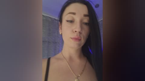 A video of a young woman with long black hair, fair skin, and dark eyes, wearing a black spaghetti-strap top and gold necklace, standing indoors with a blue-tinted wall behind her.