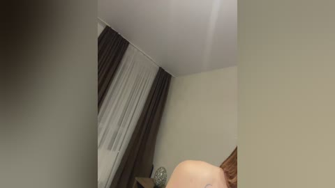 Media: A video of a dimly lit bedroom with a beige wall, a white ceiling, and dark brown curtains partially drawn. A bald man with a light skin tone, wearing a white T-shirt, is partially visible in the lower right corner.