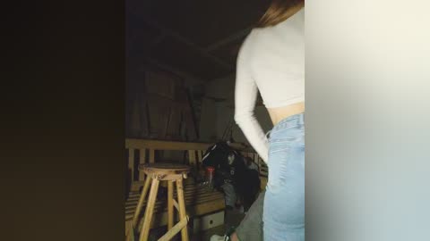 Video of a young woman with long, light brown hair, wearing a white crop top and high-waisted light blue jeans, standing in a dimly lit workshop with wooden stools, tools, and cluttered shelves in the background.