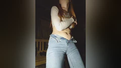 Video of a young woman with long, straight red hair, wearing a beige crop top and light blue jeans. She stands in a dimly lit room with a wooden crib in the background.