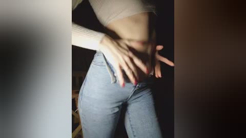 Video of a person with fair skin, wearing a light-colored, long-sleeved top and tight, light blue jeans, standing in a dimly lit room with a wooden chair in the background.