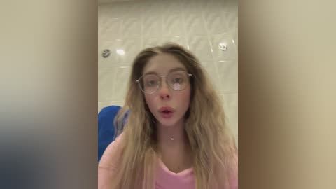 A video of a young Caucasian woman with long, wavy blonde hair, wearing glasses and a pink shirt, in a bathroom with white tiled walls and a blue towel.