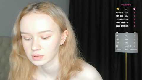 Media: Video of a fair-skinned, blonde woman with closed eyes, wearing minimal makeup, standing against a dark curtain. A camera overlay displays exposure settings and shutter speed.