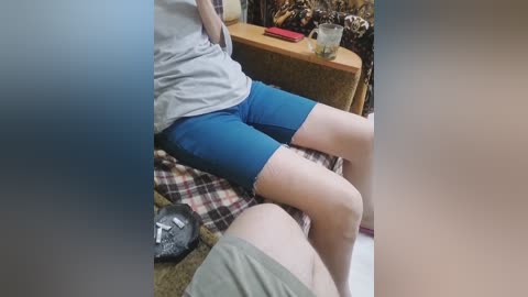 Video of a person in blue shorts and gray shirt, seated on a patterned couch, with a red item and shoes on the floor.