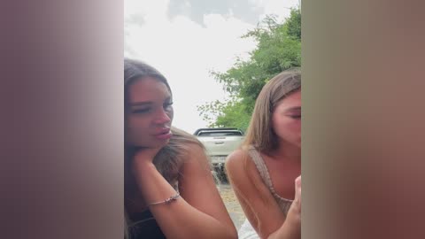 Media: Video of two young women, one with long dark hair, the other with straight blonde hair, peering through a narrow opening. They are outdoors, next to a parked car, under a cloudy sky.