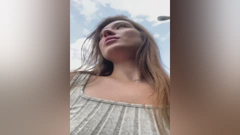 Media: Video of a young woman with long brown hair, wearing a white ribbed tank top, looking upward at a cloudy sky.