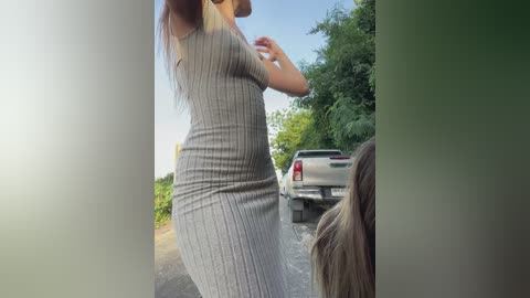 Media: Video of a woman in a gray, ribbed dress standing on a country road. She has long, light brown hair and is partially obscured by a car and foliage.