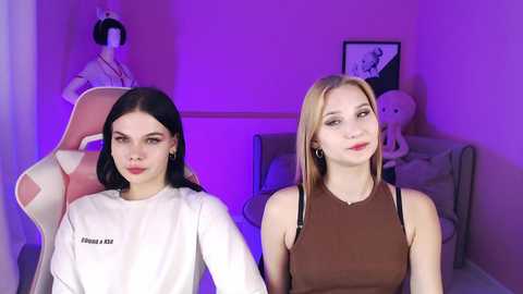 Video of two young women with fair skin and straight hair, one in a white shirt, the other in a brown tank top, standing against a purple-lit room with plush toys and a doll.