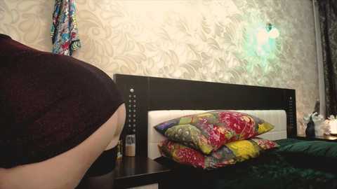A video of a woman in a dark shirt bending over a bed with colorful floral pillows and a green comforter. The room features a white, patterned wallpaper and a blue-green light fixture.