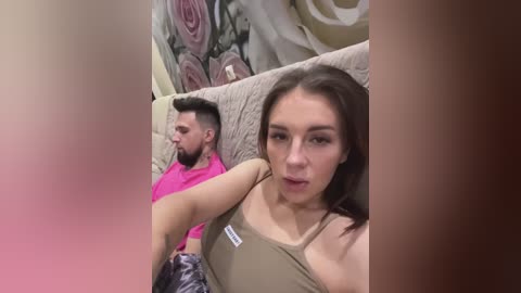 Media: Video of a young woman with light skin and straight brown hair, wearing a beige tank top, taking a selfie. Behind her, a man with a beard, wearing a pink shirt, sits on a beige couch with floral wallpaper in the background.