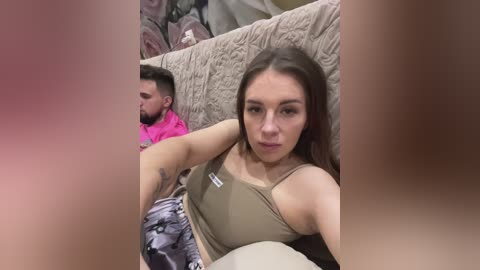 Media: Video of a woman with long brown hair, wearing a beige sports bra and patterned shorts, seated on a beige couch. A man with short dark hair and a beard, wearing a pink shirt, sits behind her. Background features floral-patterned pillows.