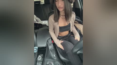 Media: Video of a young woman with long dark hair, wearing a black tube top and black pants, sitting in a car, looking down.