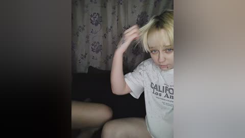 Media: Video of a young, blonde-haired girl with a worried expression, wearing a white t-shirt with \"Californians for Trump\" text. She sits in a dimly lit room with floral-patterned curtains and a black cushion in the background.