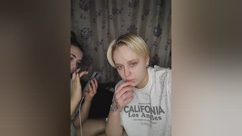 Media: Video of a blonde woman with a tattoo on her right hand, wearing a \"California Los Angeles\" shirt, taking a selfie with a phone, partially obscured by a curtain.
