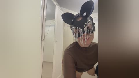 Video of a person wearing a black, glittery rabbit mask and a brown shirt, kneeling on beige carpet in a dimly lit room with white walls and a door.