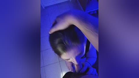 A video showing a person with short dark hair kneeling on a tiled floor, their head resting on another person's hand under a blue light.