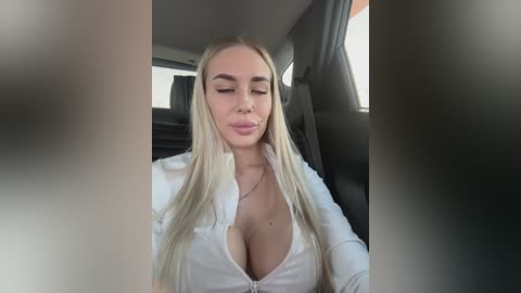 Video of a blonde woman with long hair, wearing a low-cut white shirt, sitting in a car, her cleavage visible.