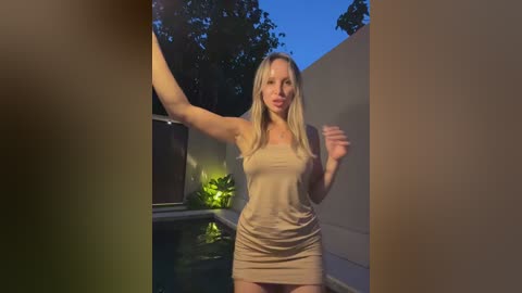Media: A video of a blonde woman in a beige mini dress standing by an outdoor pool at dusk, with trees and a white wall in the background.