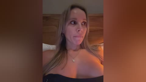 Media: A video of a woman with long, straight, blonde hair, wearing a black off-shoulder top, sitting on a bed with wooden headboard.