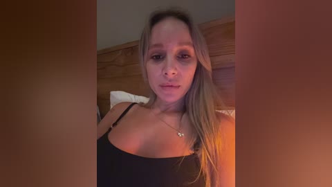 Media: Video of a light-skinned woman with long blonde hair, wearing a black spaghetti-strap top, sitting on a bed with a wooden headboard. She has a neutral expression.