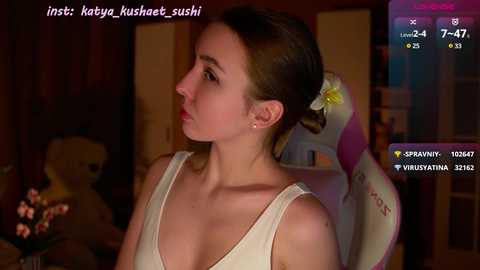 Media: Video of a young woman with light skin and brown hair in a white tank top, sitting in a pink gaming chair in a dimly lit room.