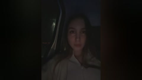 Media: Video of a young woman with dark hair, wearing a white blouse, seated in a dimly lit car. The background is dark, creating a moody atmosphere.