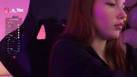 Video of a young woman with long red hair, wearing a black top, sitting in a black gaming chair under purple lighting.