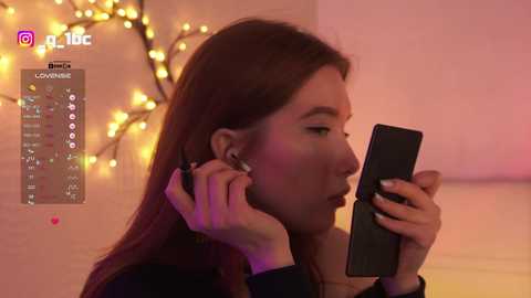Video of a young woman with long brown hair, wearing headphones, focused on her smartphone, in a cozy room with warm, yellow string lights in the background.